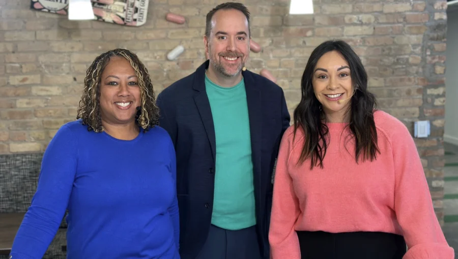 Three people stand indoors in front of a brick wall, smiling at the camera. The person on the left wears a blue top, the middle person wears a green shirt and dark blazer, and the right person wears a pink top.